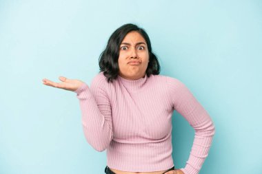 Genç Latin bir kadın, mavi arka planda izole edilmiş. Sorgulama sırasında omuz silkiyor ve şüphe ediyor..