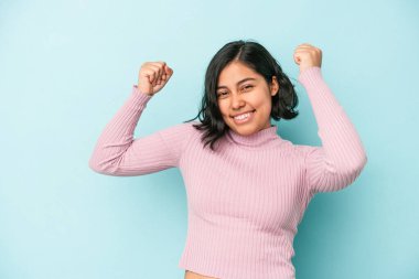 Genç Latin bir kadın mavi arka planda izole edilmiş tasasız ve heyecanlı tezahüratlar yapıyor. Zafer konsepti.