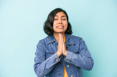 Mavi arka planda izole edilmiş genç Latin bir kadın. El ele tutuşup ağzının kenarında dua ediyor. Kendine güveniyor..