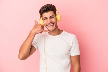 Pembe arka planda izole edilmiş müzik dinleyen genç beyaz adam parmaklarıyla bir cep telefonu çağrısı hareketi gösteriyor..
