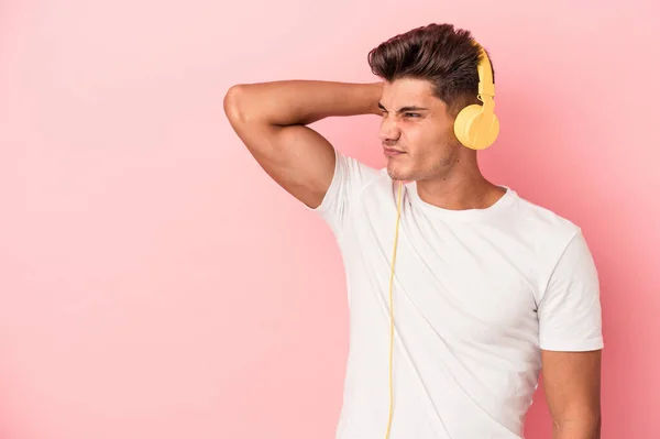Young caucasian man listening to music isolated on pink background touching back of head, thinking and making a choice.