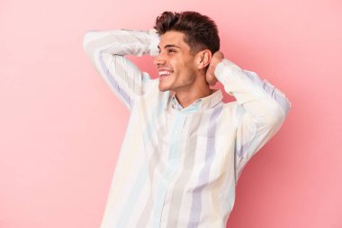 Pembe arka planda izole edilmiş genç beyaz bir adam kendini güvende hissediyor, elleri başının arkasında..