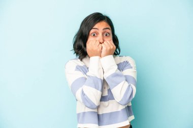 Mavi arka planda izole edilmiş, tırnak yiyen, gergin ve endişeli genç bir Latin kadın..