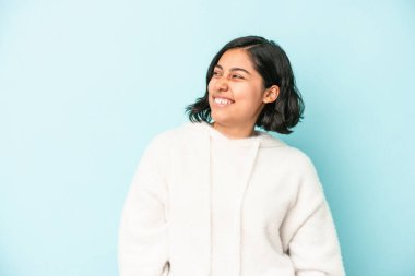 Mavi arka planda izole edilmiş genç Latin bir kadın gevşemiş ve mutlu bir şekilde gülüyor, boynunda dişlerini gösteriyor..