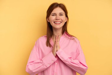 Sarı arka planda izole edilmiş genç bir İngiliz kadın el ele tutuşup ağzının kenarında dua ediyor, kendinden emin hissediyor..