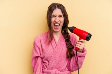 Sarı arka planda izole edilmiş saç kurutma makinesi tutan genç bir kadın çok sinirli ve agresif bağırıyor..
