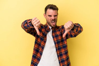 Sarı arka planda izole edilmiş genç beyaz adam baş parmağını aşağı gösteriyor ve hoşlanmadığını ifade ediyor..