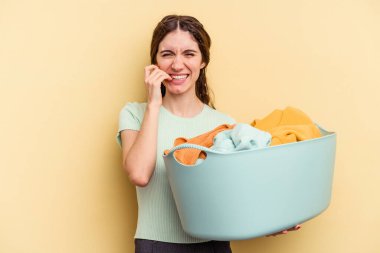 Sarı arka planda izole edilmiş, tırnak yiyen, gergin ve endişeli bir çamaşır sepeti tutan beyaz bir kadın..