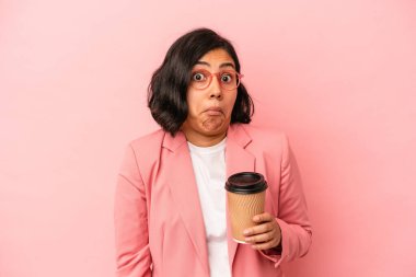 Elinde kahve tutan genç Latin kadın pembe arka planda izole edilmiş omuzlarını silkeliyor ve gözleri açık..
