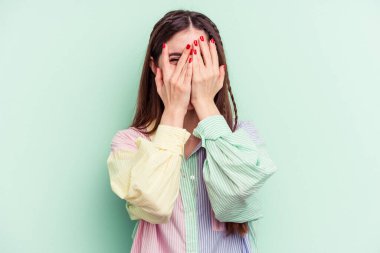 Yeşil arka planda izole edilmiş genç beyaz kadın kameraya parmaklarıyla göz kırpıyor, yüzünü kapatmaktan utanıyor..