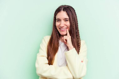 Genç beyaz kadın yeşil arka planda izole edilmiş mutlu ve kendinden emin gülümsüyor, elleriyle çenesine dokunuyor..