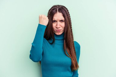 Yeşil arka planda izole edilmiş genç beyaz kadın yumruğunu kameraya gösteriyor, agresif yüz ifadesi var..