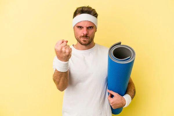 年轻的体育白人男子去上瑜伽课 同时拿着一个被黄色背景隔开的垫子 用拳头对着镜头 露出咄咄逼人的表情 — 图库照片