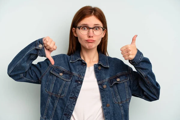 Mavi arka planda izole edilmiş genç bir İngiliz kadın baş parmağını kaldırıyor ve baş parmağıyla işaret ediyor. Zor seçim kavramı.