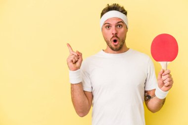 Sarı arka planda pinpon raketi tutan genç beyaz adam yan tarafı işaret ediyor.
