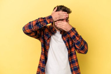Sarı arka planda izole edilmiş genç beyaz adam kameraya parmaklarıyla göz kırpıyor, yüzünü kapatmaktan utanıyor..