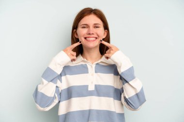 Mavi arka planda izole edilmiş genç bir İngiliz kadın gülümsüyor, parmaklarını ağıza doğrultuyor..