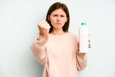 Mavi arka planda izole edilmiş bir şişe süt tutan genç İngiliz kadın yumruğunu kameraya gösteriyor, agresif yüz ifadesi..