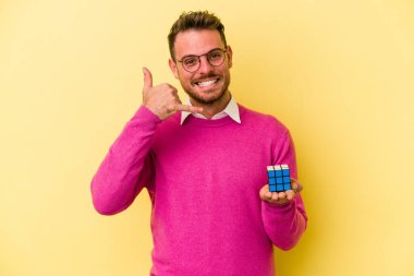 Arka planda Rubiks küpünü tutan genç beyaz adam parmaklarıyla bir cep telefonu hareketi gösteriyor..
