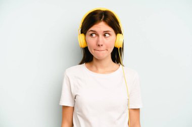 Mavi arka planda izole edilmiş müzik dinleyen genç İngiliz kadın kafası karışmış, kuşkulu ve kendinden emin değil..