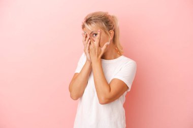 Pembe arka planda izole edilmiş genç Rus kadın parmaklarının arasından korkmuş ve gergin bir şekilde göz kırpıyor..