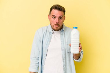 Sarı arka planda izole edilmiş bir şişe süt tutan beyaz bir adam omuzlarını silker ve gözleri açık kalır..