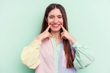 Yeşil arka planda izole edilmiş genç beyaz kadın gülümsüyor, parmaklarını ağzına doğrultuyor..