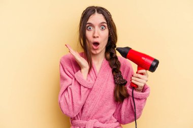 Sarı arka planda izole edilmiş saç kurutma makinesini tutan genç bir kadın şaşırdı ve şok oldu..