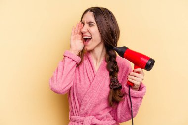 Sarı arka planda izole edilmiş saç kurutma makinesini tutan genç bir kadın bağırıyor ve avuç içini açık tutuyor..