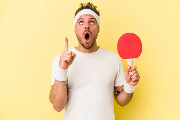 年轻的高加索人手持乒乓球球拍 隔离在黄色背景下 指向上方 张开嘴 — 图库照片