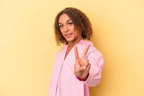 若いラテン語の性転換女性は黄色の背景に孤立喜びと屈託のない指で平和のシンボルを示す — ストック写真