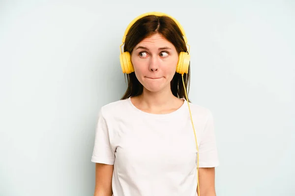 Young English woman listening to music isolated on blue background confused, feels doubtful and unsure.