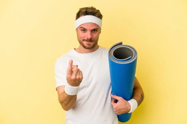 年轻的体育白人男子去上瑜伽课 同时拿着一个被黄色背景隔开的垫子 用手指指著你 好像邀请你走近些 — 图库照片
