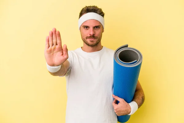 年轻的体育白人男子去上瑜伽课 同时拿着一个被黄色背景隔离的垫子 手伸展开 显示出停止的迹象 阻止了你 — 图库照片