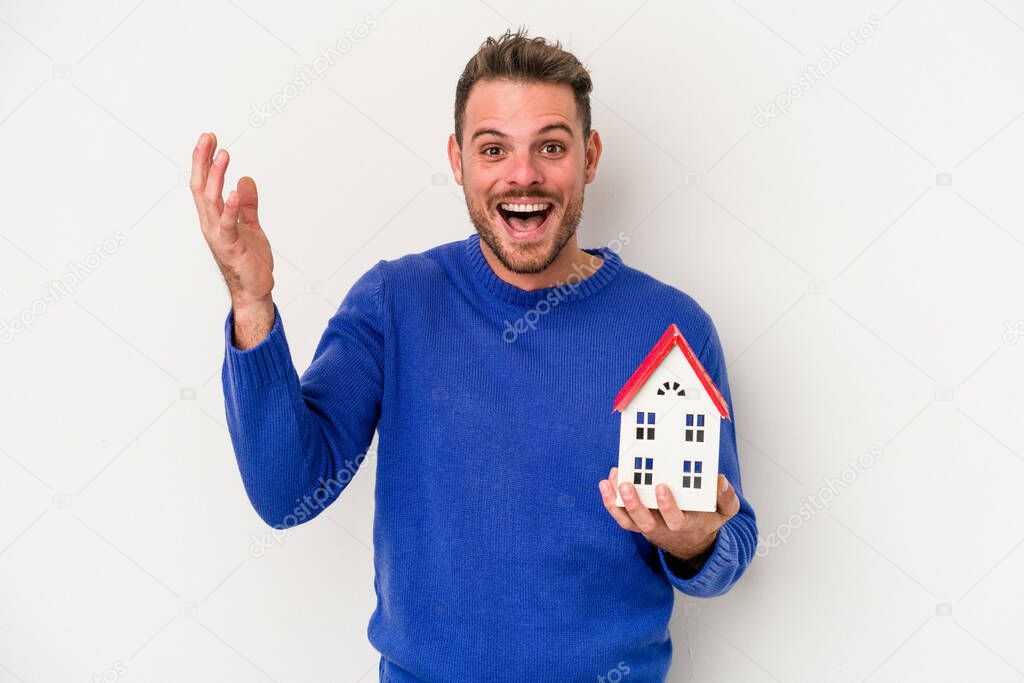 Young caucasian man holding a little house isolated on white background receiving a pleasant surprise, excited and raising hands.