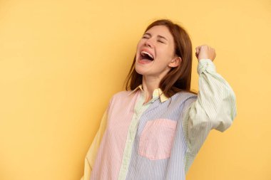 Sarı arka planda izole edilmiş genç İngiliz kadın zaferi, tutkuyu ve coşkuyu, mutlu ifadeyi kutluyor..