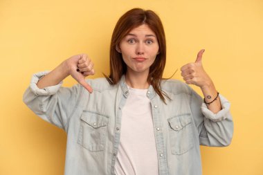 Sarı arka planda izole edilmiş genç bir İngiliz kadın baş parmağını kaldırıyor ve baş parmağıyla işaret ediyor. Zor seçim kavramı.