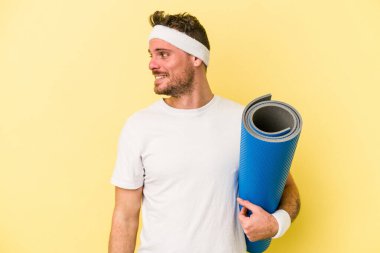 Sarı arka planda izole edilmiş bir minderi tutarken yoga derslerine giden genç beyaz adam gülümsüyor, neşeli ve keyifli görünüyor..