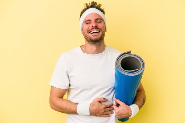 Sarı arka planda izole edilmiş bir minderi tutarken yoga derslerine giden genç beyaz bir adam gülüyor ve eğleniyor..