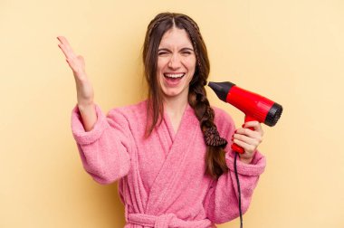 Sarı arka planda izole edilmiş saç kurutma makinesini tutan genç bir kadın hoş bir sürpriz bekliyor, heyecanlı ve el kaldırıyor..
