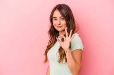 Pembe arka planda izole edilmiş genç beyaz kadın bir göz kırpıyor ve elinde iyi bir el hareketi tutuyor..