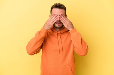 Sarı arka planda izole edilmiş genç beyaz adam elleriyle gözlerini kapatmaktan korkuyor..
