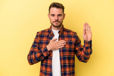 Sarı arka planda izole edilmiş genç beyaz adam yemin ediyor, elini göğsüne koyuyor..