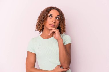 Genç Latin transseksüel kadın pembe arka planda izole edilmiş düşünüyor ve yukarı bakıyor, yansıtıyor, düşünüyor, hayal kuruyor..