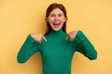Sarı arka planda izole edilmiş genç bir İngiliz kadın parmağını göstererek şaşırdı, geniş bir gülümsemeyle..