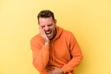 Sarı arka planda izole edilmiş genç beyaz adam mutlu bir şekilde gülüyor ve ellerini karnının üstünde tutarak eğleniyor..