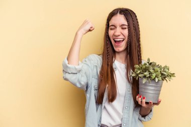Sarı arka planda izole edilmiş bir bitkiyi elinde tutan beyaz bir kadın zaferden sonra yumruğunu kaldırıyor, kazanan konsept..