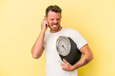 Kafkasyalı genç adam sarı arka planda izole edilmiş bir tartı tutuyor kulakları elleriyle kapatıyor..
