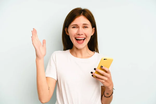 青の背景に隔離された携帯電話を持っている若い英語の女性は 楽しい驚き 興奮し 手を上げる — ストック写真