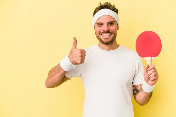 若いですCaucasian男保持Ping Pongラケット孤立した上に黄色の背景笑顔と上げ親指アップ — ストック写真